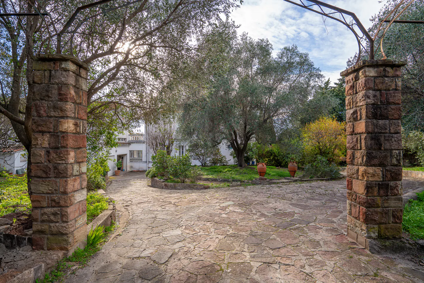 Stone pathway leading to a white house, framed by brick pillars and trees. Two terracotta pots sit on a grassy area.