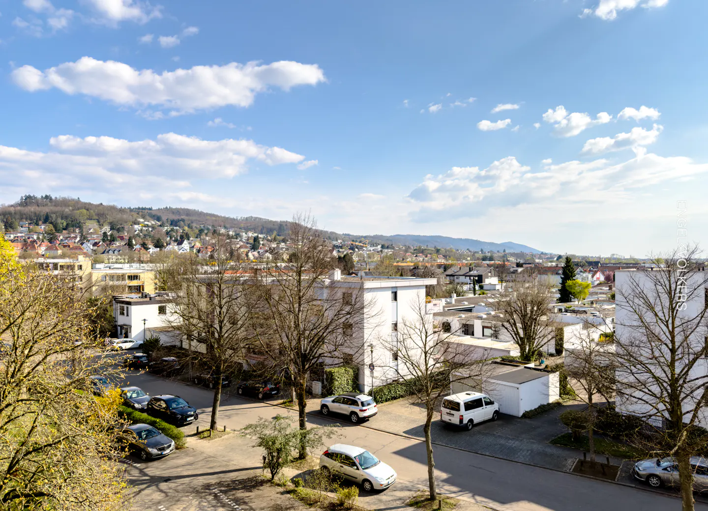 View of a European town with white buildings, cars, and a tree-covered hill under a blue sky with clouds.
