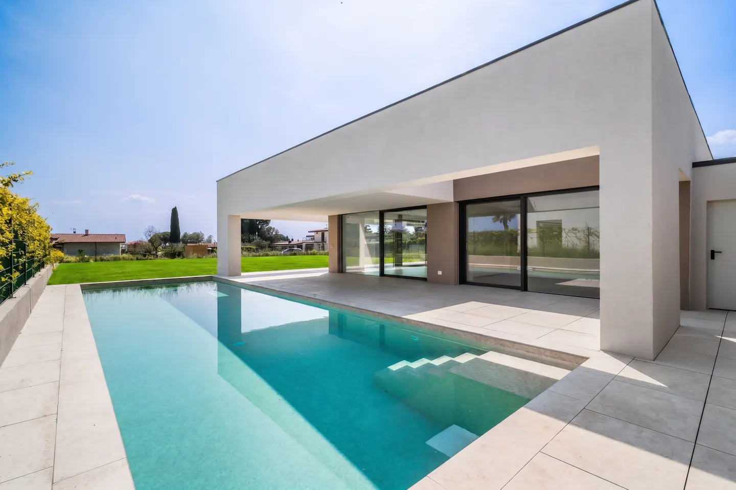 Modern white house with a turquoise pool, patio, and sliding glass doors under a sunny blue sky.