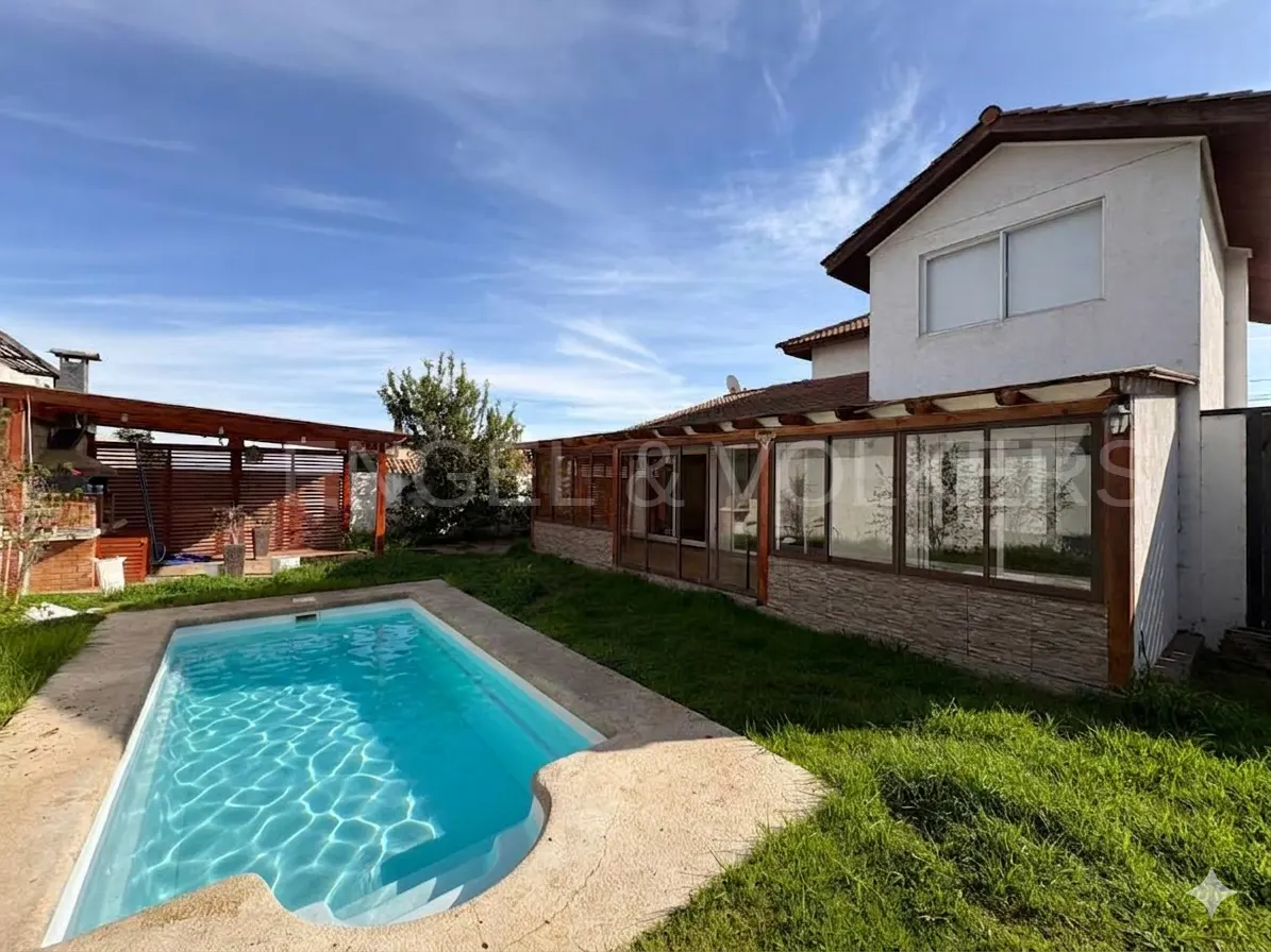 Backyard with a rectangular swimming pool, green lawn, and a white house with a wooden patio cover under a blue sky.