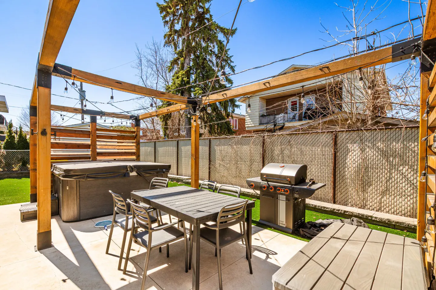 Outdoor patio with a hot tub, grill, and dining table under a wooden pergola with string lights. A chain-link fence surrounds the yard.