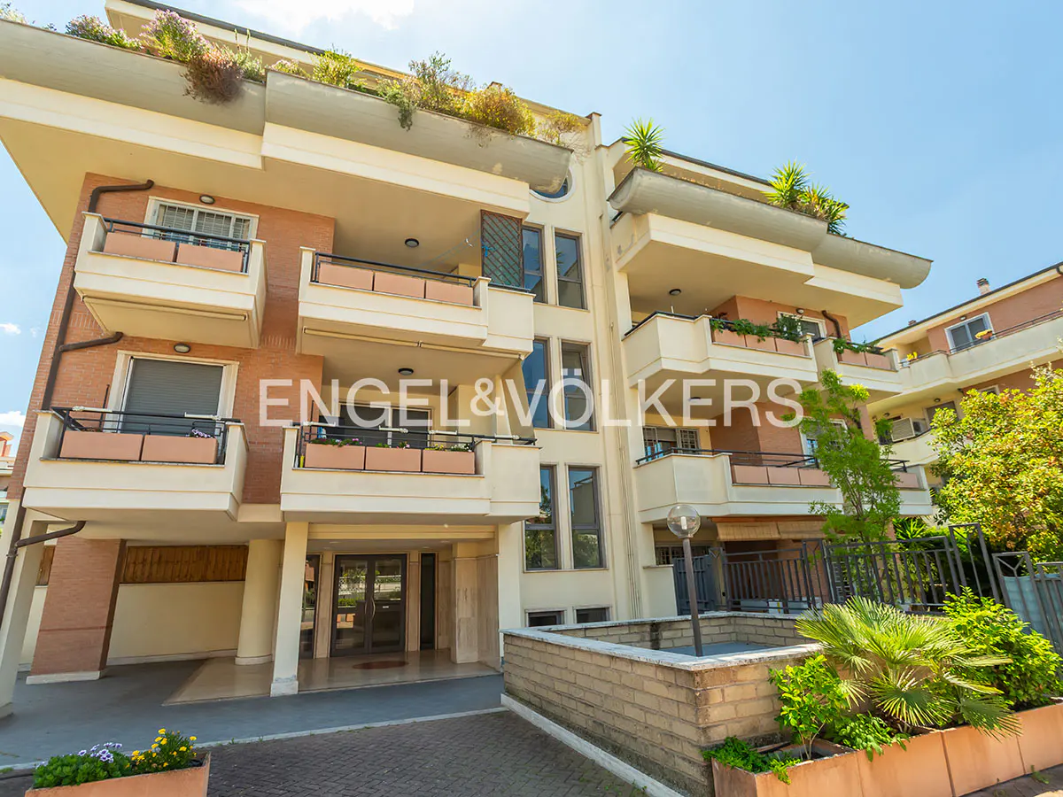 Exterior of a multi-story apartment building with balconies and the Engel & Völkers logo. The building is brick and beige with green plants.