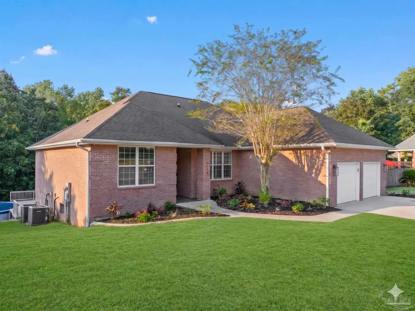 Brick house with a dark roof, green lawn, and a tree in the front yard under a blue sky.