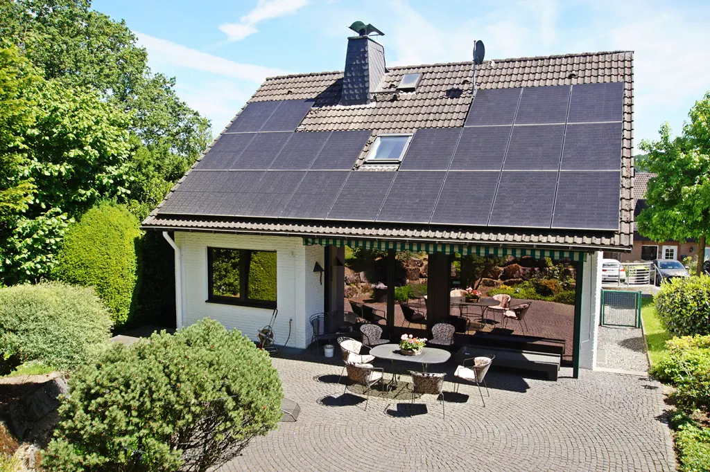 White house with solar panels on a tiled roof, featuring a paved patio with outdoor furniture and lush green surroundings.