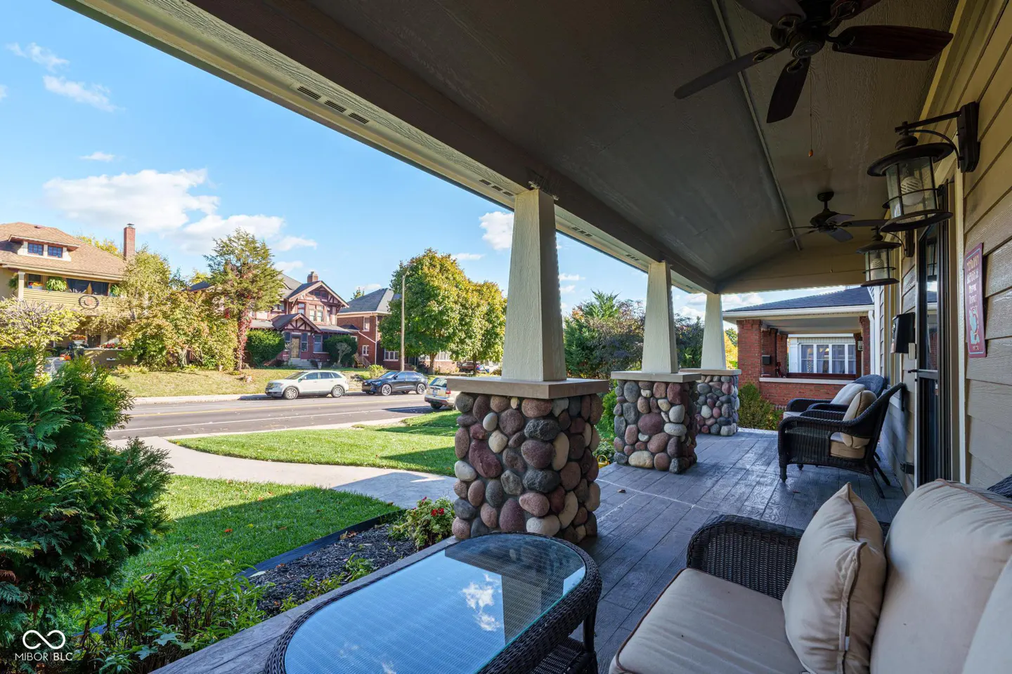 Covered porch with wicker furniture, stone pillars, and ceiling fans. Street view with houses and cars in the background.