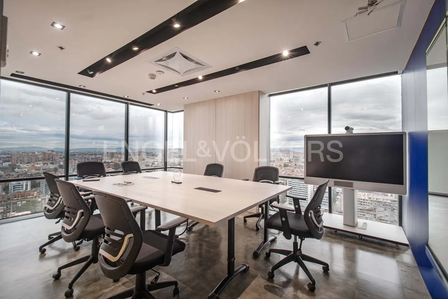 Bright conference room with a long table, black chairs, and a large screen. Windows offer a city view. Engel & Völkers logo visible.