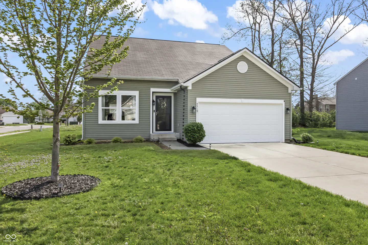 Exterior of a one-story, light green house with a white garage door and a black front door. A tree stands in the front yard.