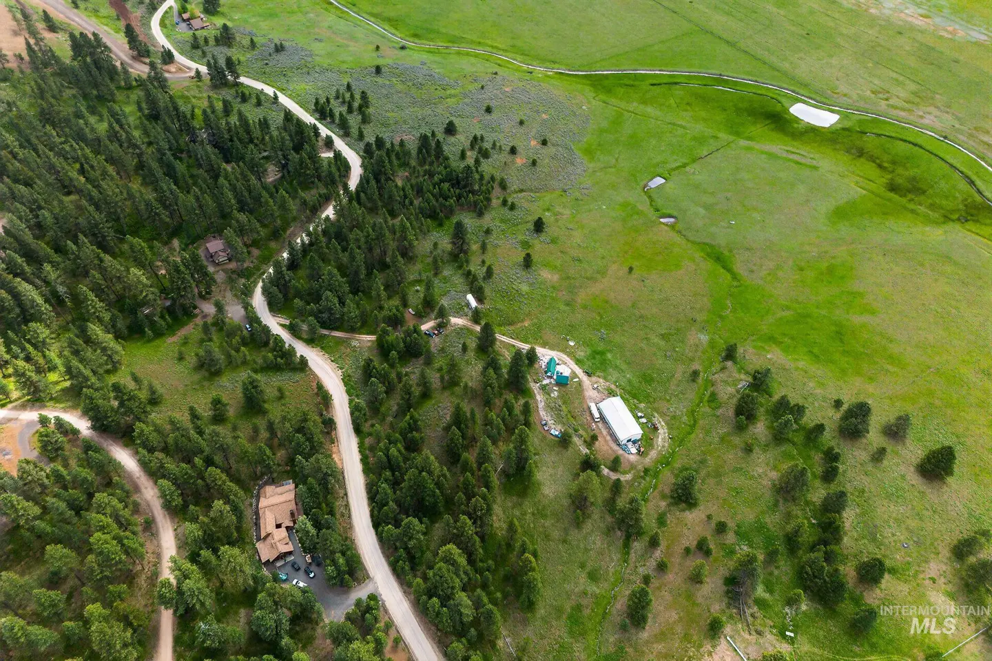 Aerial view of a property with a winding road, green meadows, and dense pine forests. Buildings are scattered throughout the landscape.