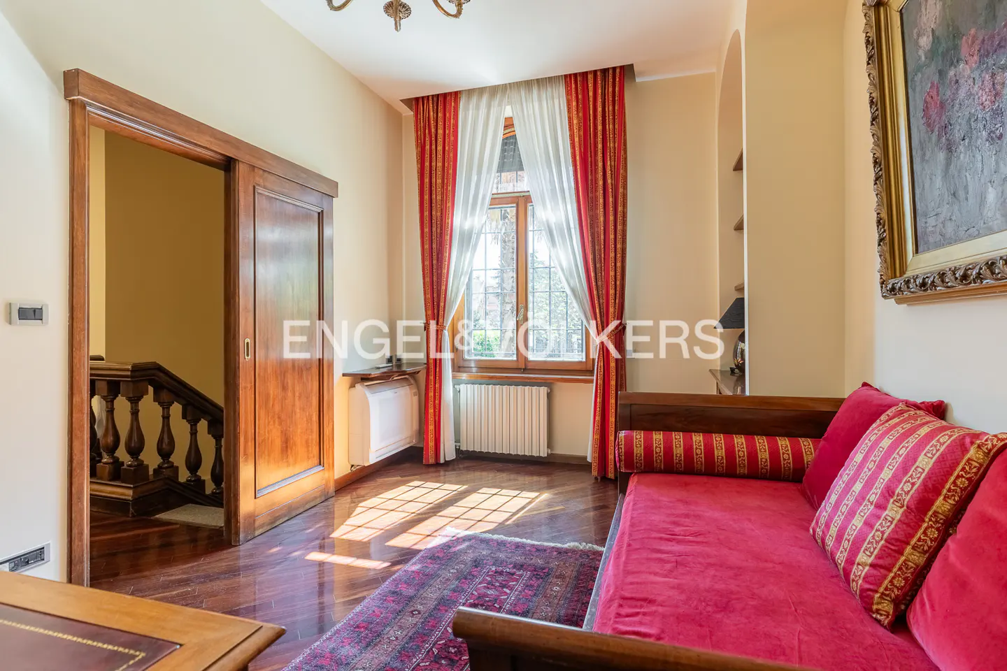 A sunlit room with a red sofa, a window with red curtains, and a wooden sliding door leading to a staircase.
