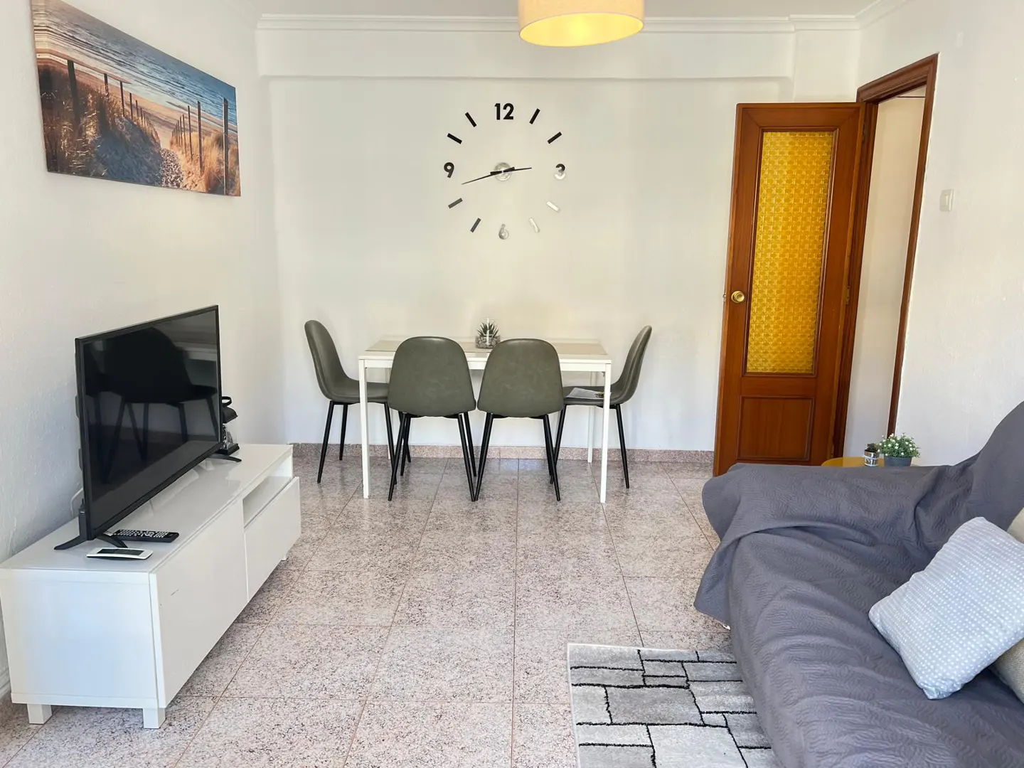 Living room with a white TV stand, dining table with four grey chairs, wall clock, and a grey sofa on tiled flooring.