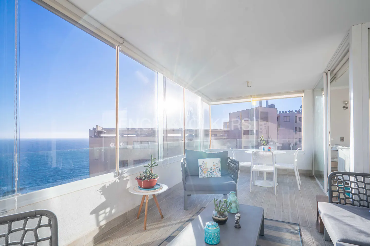 Bright sunroom with ocean view. Gray chairs and table sit on a wood floor. Glass walls overlook the blue ocean and sky.