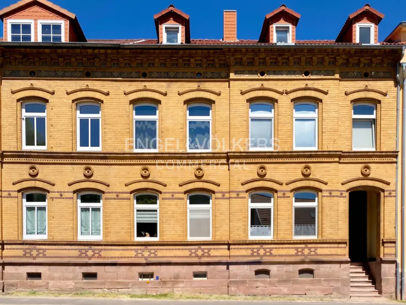 Exterior view of a historic yellow brick building with arched windows, a stone base, and four dormers under a blue sky.