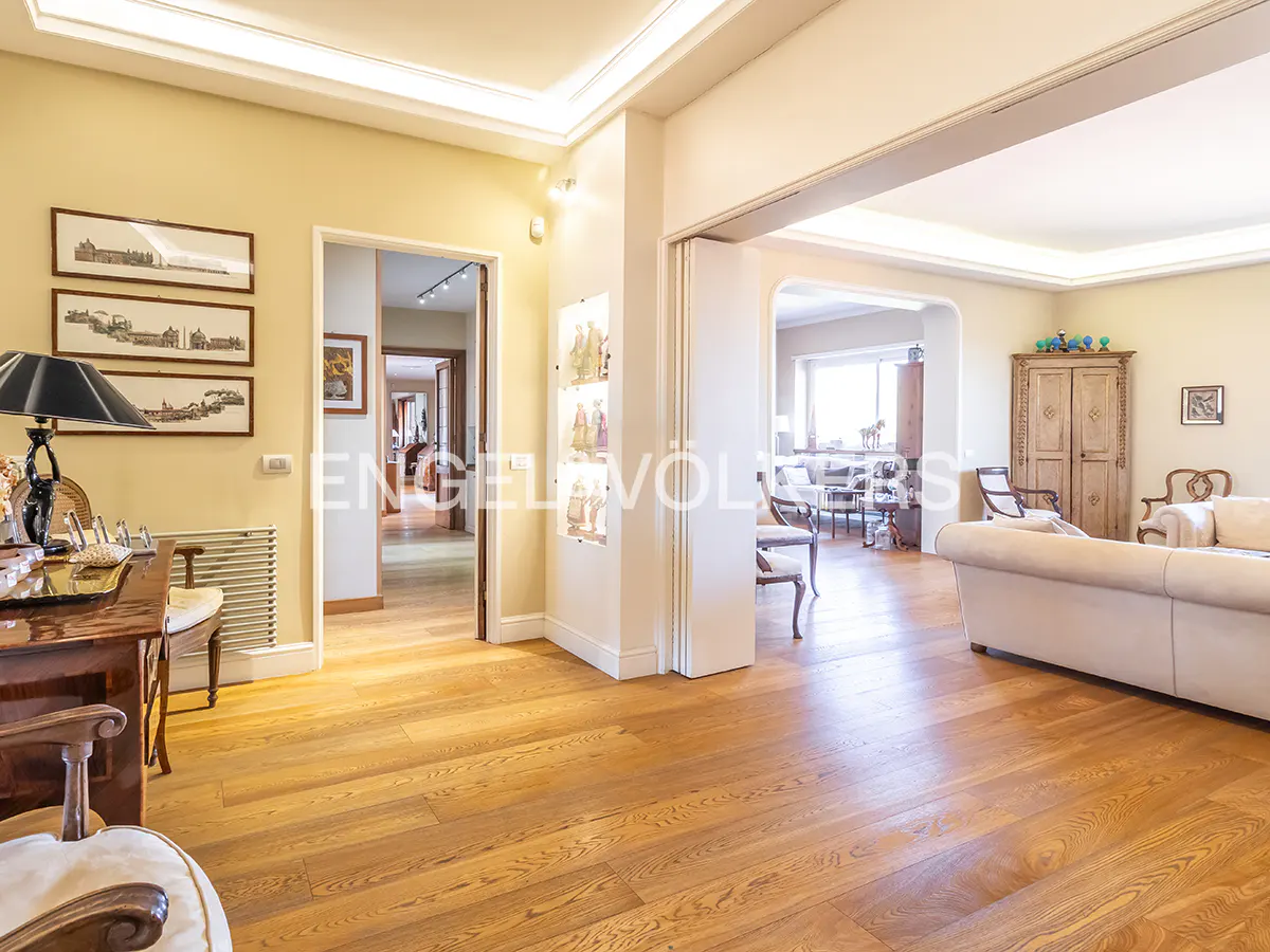 A bright, open-concept living space with hardwood floors, cream walls, and multiple doorways. A desk and sofa are visible.