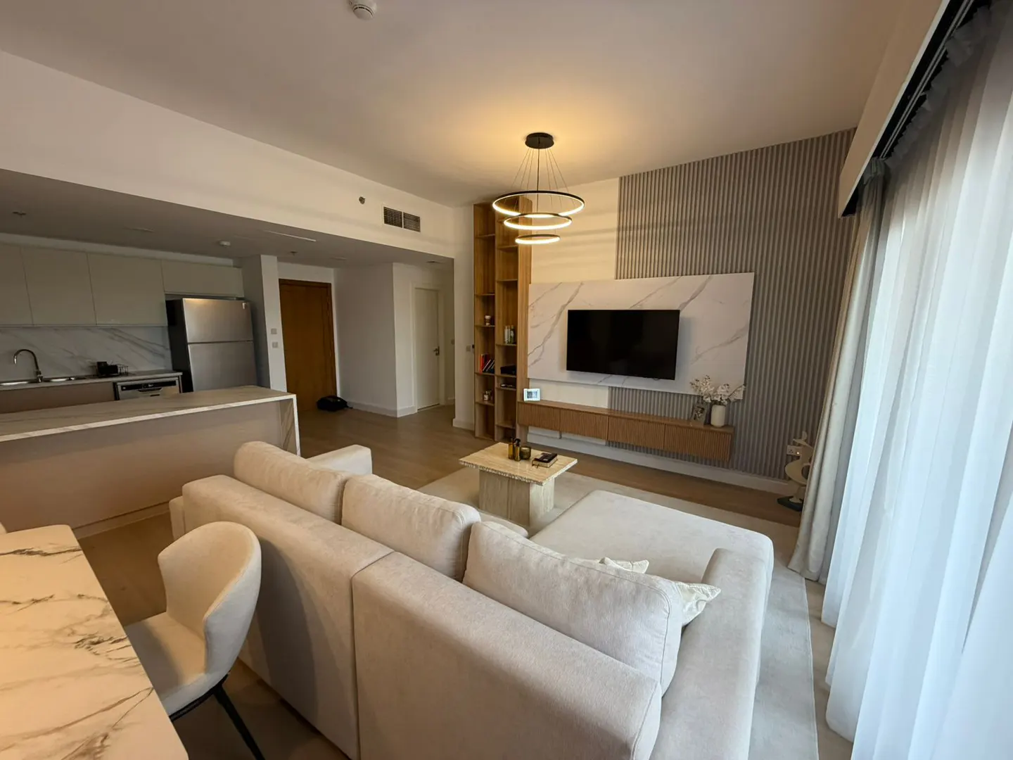 A modern living room with a beige sectional sofa, a marble coffee table, and a TV on a marble wall. A kitchen is visible in the background.