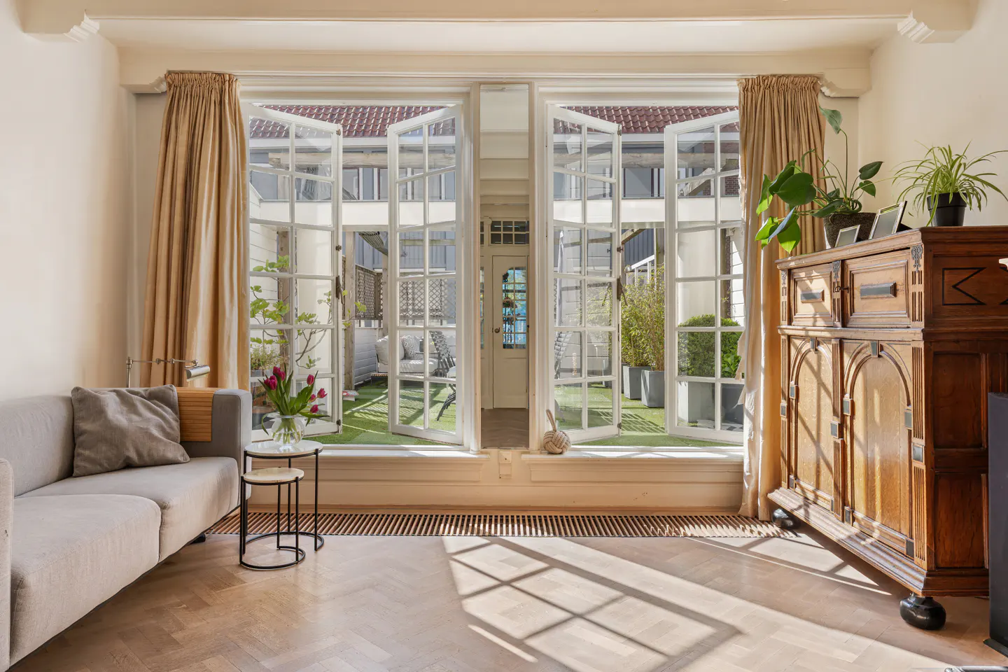 Living room with open French doors to a courtyard. A gray sofa, wood floors, and a wooden cabinet are visible.