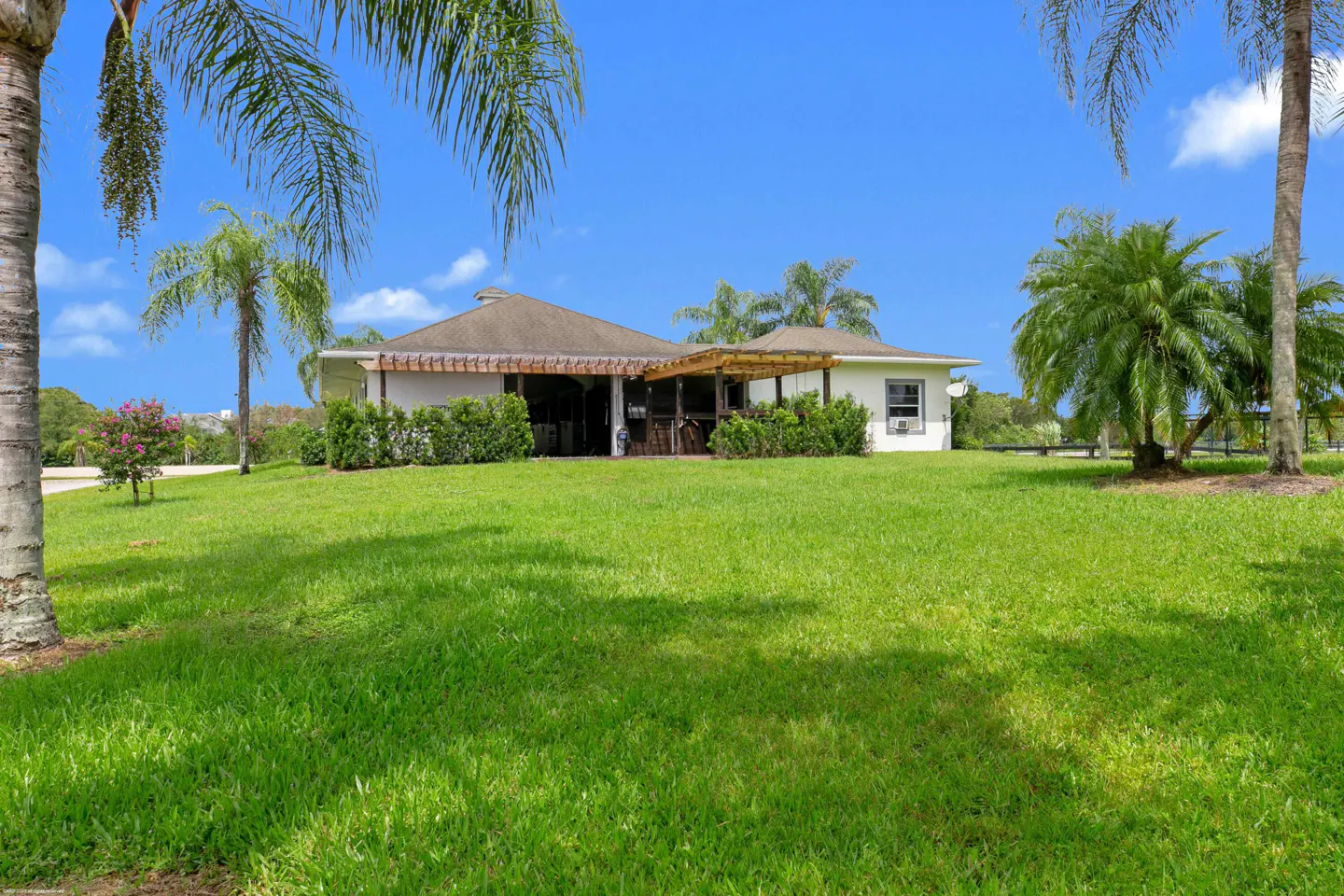 A white house with a brown roof and pergola sits on a green lawn under a blue sky with palm trees.