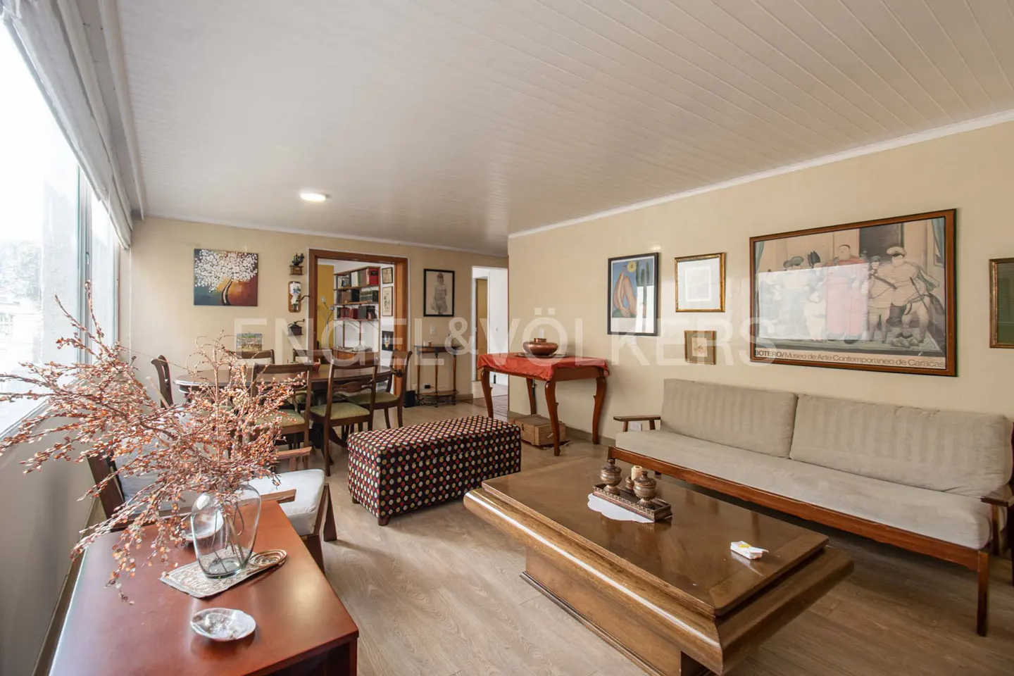 A living room with beige walls, wood floors, a beige sofa, and a wooden coffee table. Artwork hangs on the walls.