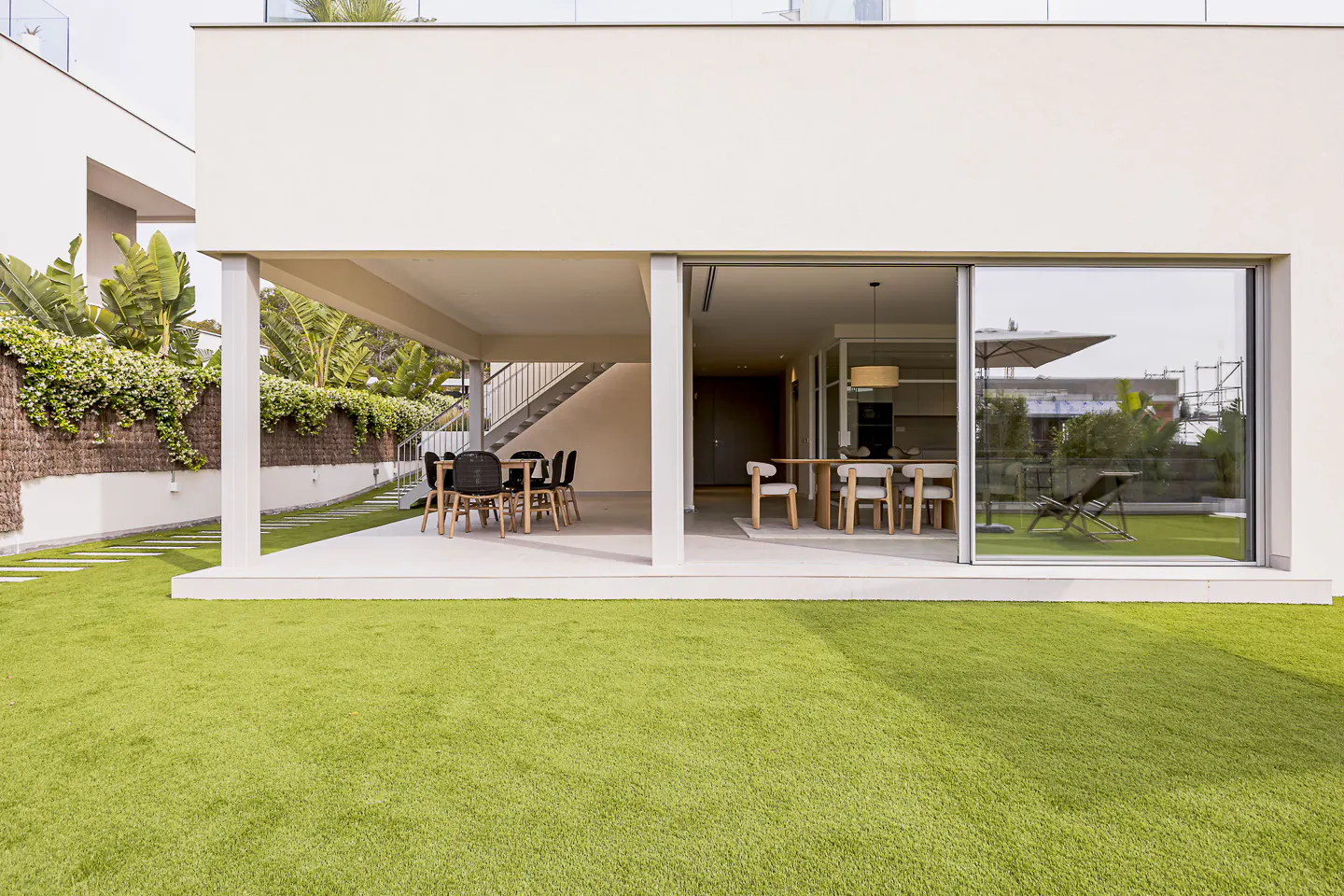 Modern home exterior with a green lawn, patio with table and chairs, and a large sliding glass door to the dining area.
