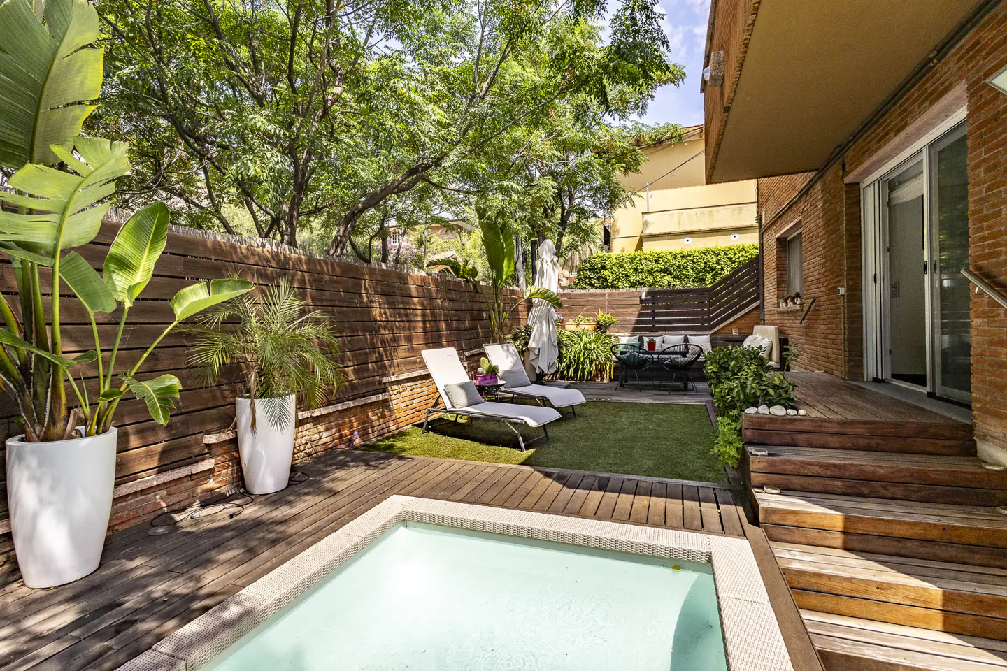 A backyard with a small pool, wooden deck, lounge chairs, and green grass. A wooden fence and trees provide privacy.