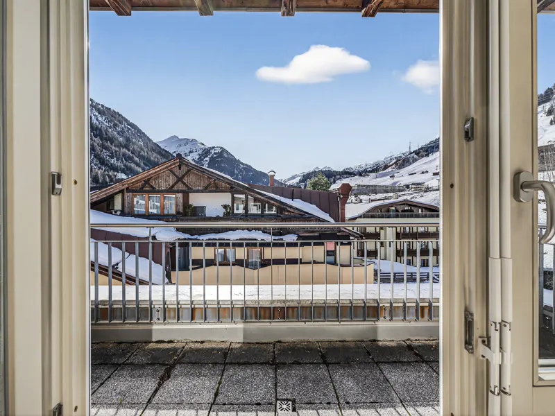 View from open balcony doors to snow-covered rooftops and mountains under a blue sky.