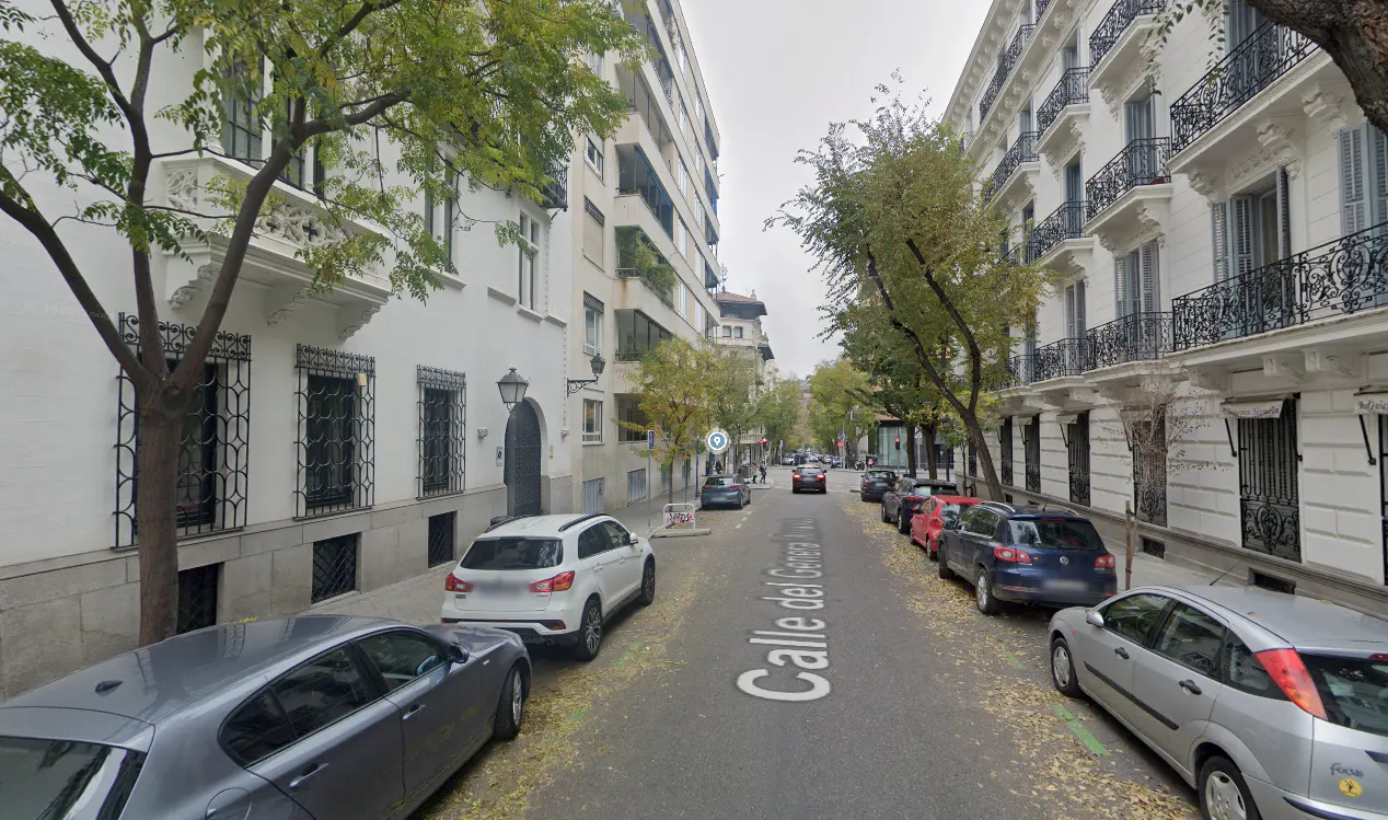 Street view of Calle del General in Madrid, Spain. Cars are parked along the sides of the street. Buildings line the street.