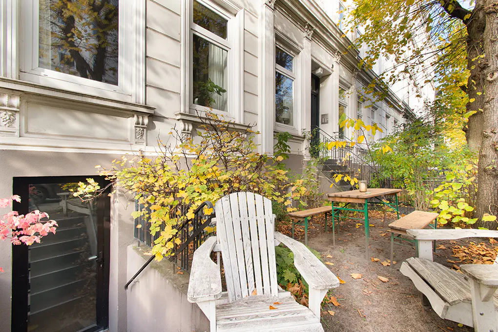 White wooden chairs and a picnic table sit in a garden area in front of a classic white building with large windows.