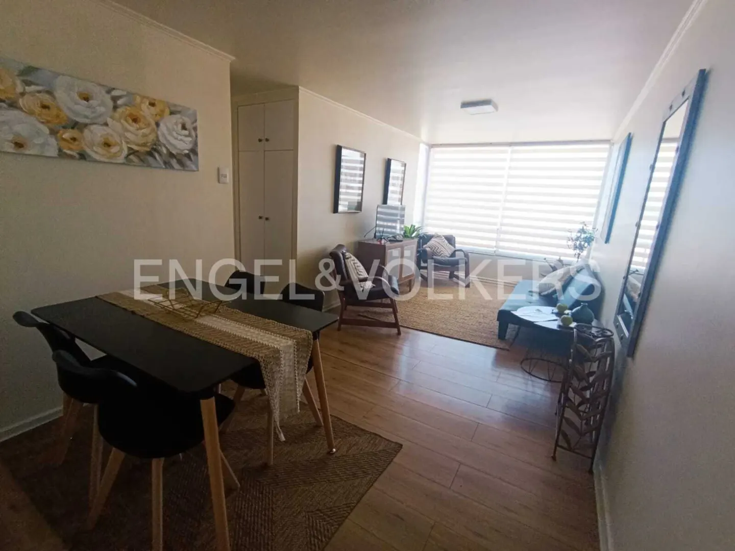 A bright apartment interior with a dining table, chairs, and a living area with a sofa and armchairs.