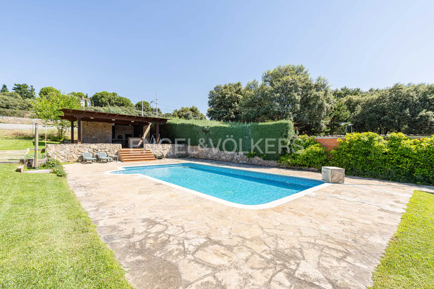 A backyard pool with a stone patio, a covered outdoor kitchen, and green trees under a blue sky.