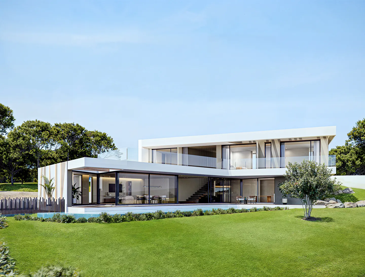 Modern white two-story house with large glass windows and doors, surrounded by green lawn and trees under a blue sky.