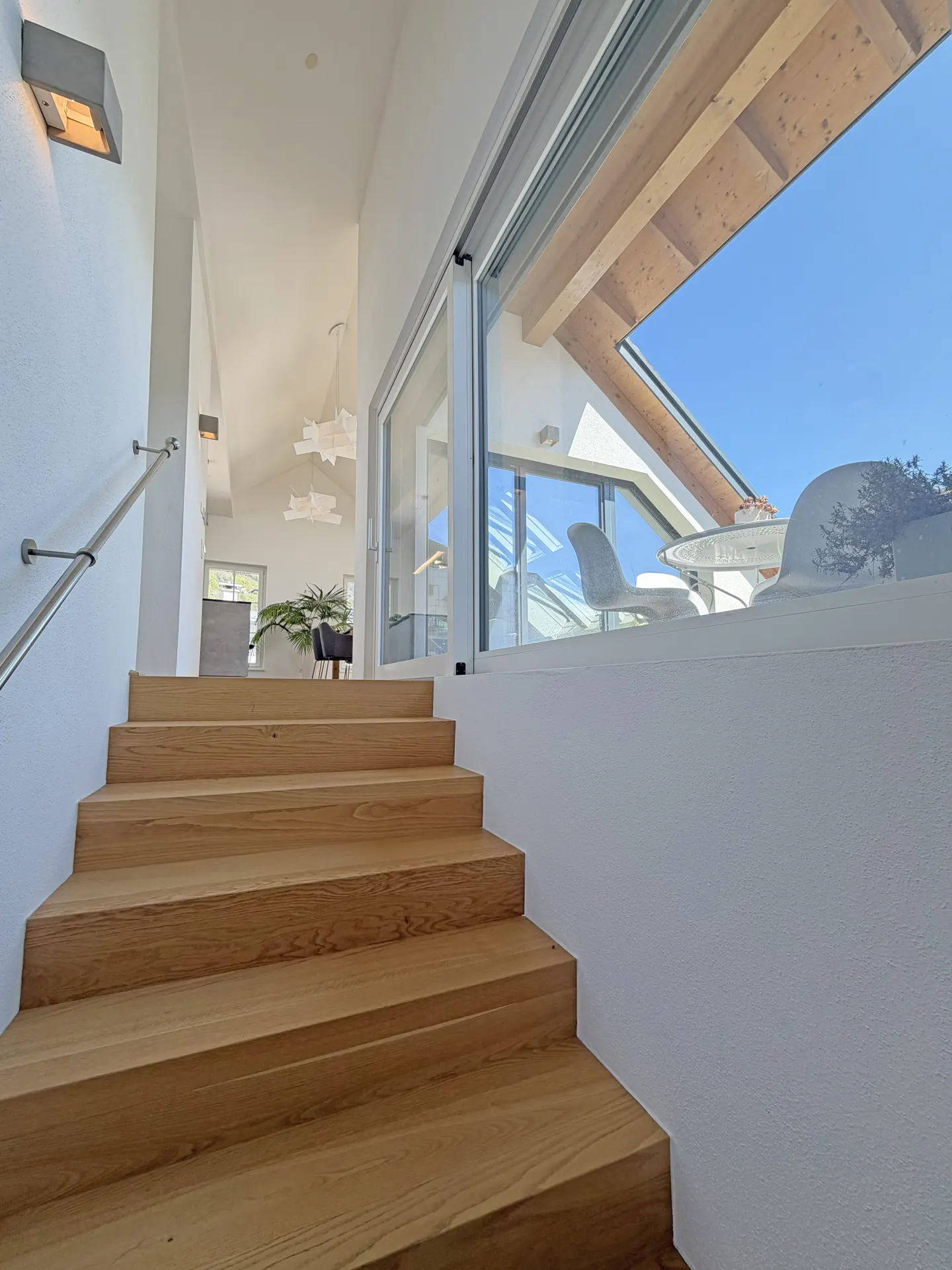 Interior view of wooden stairs with a metal handrail, leading to a bright living space with a large window and outdoor seating.