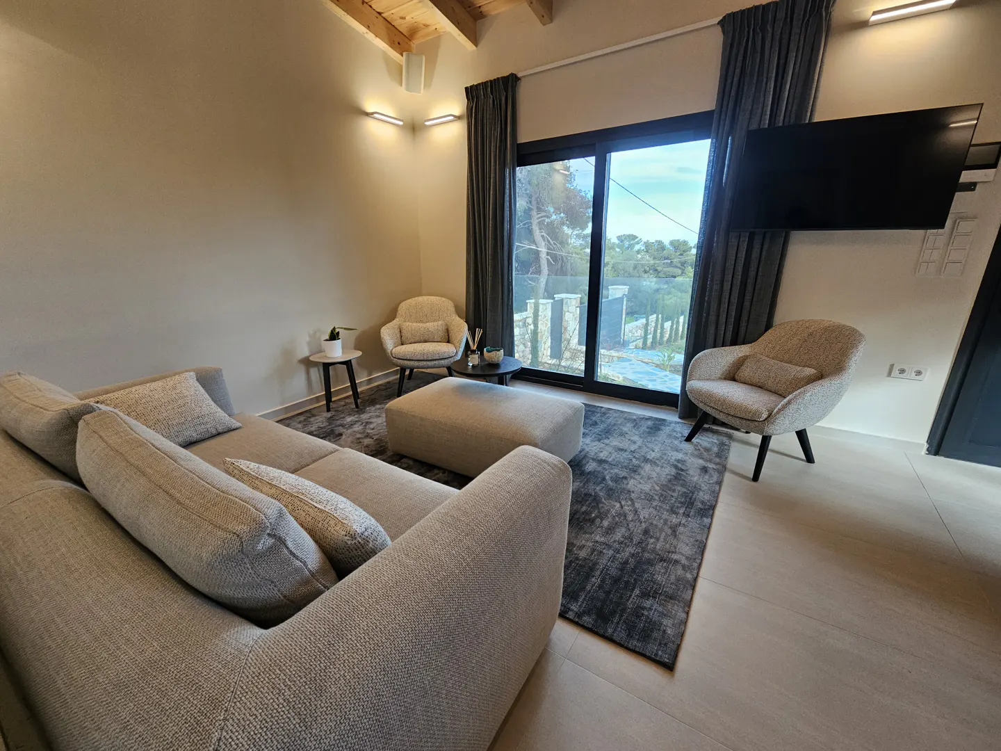 Living room with a gray sectional sofa, two armchairs, and a large window with gray curtains. A TV is mounted on the wall.