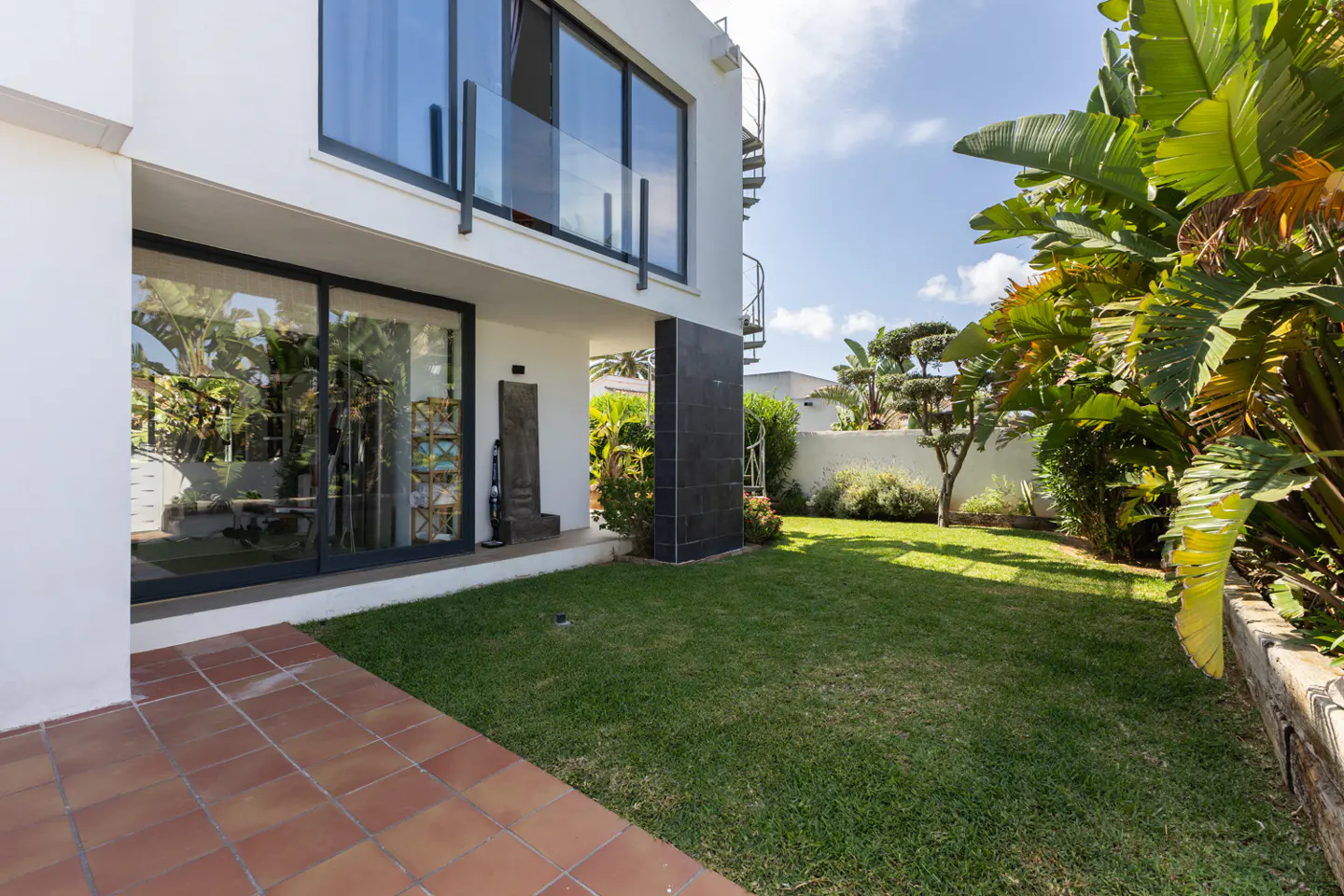 Modern white two-story home with a tiled patio, green lawn, and lush tropical plants under a bright blue sky.