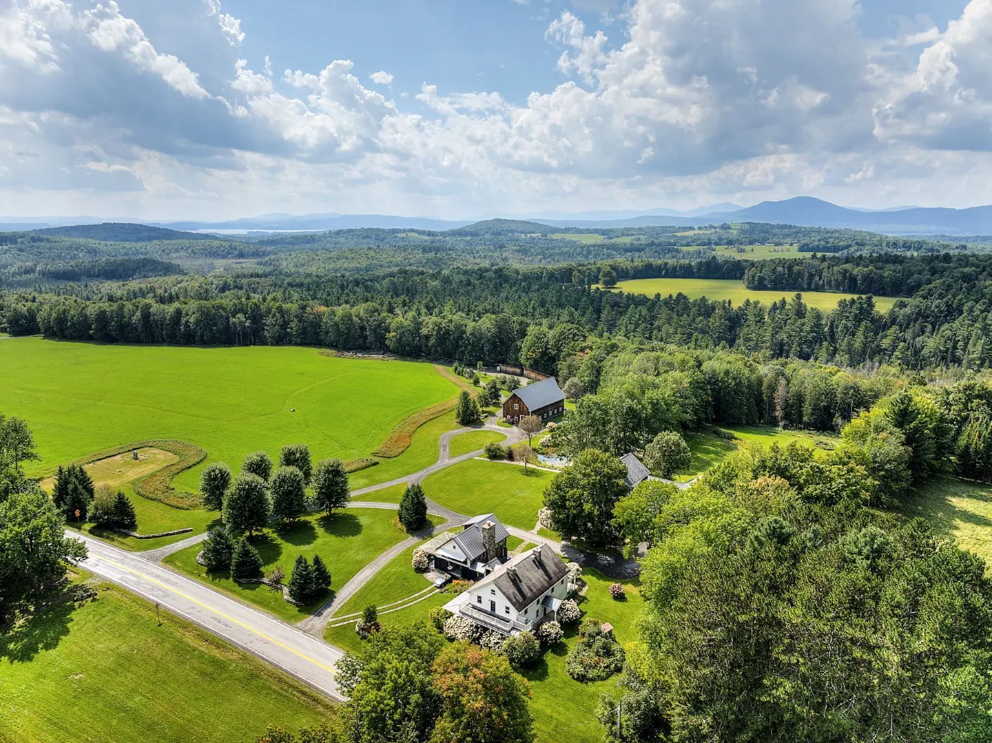 Aerial view of a white farmhouse, barn, and green fields surrounded by trees under a blue, cloudy sky.