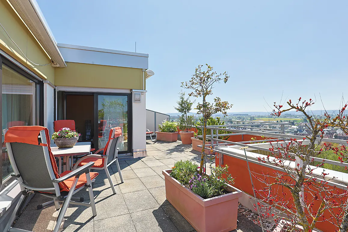 A sunny rooftop terrace with orange chairs, a table, and potted plants overlooking a town.