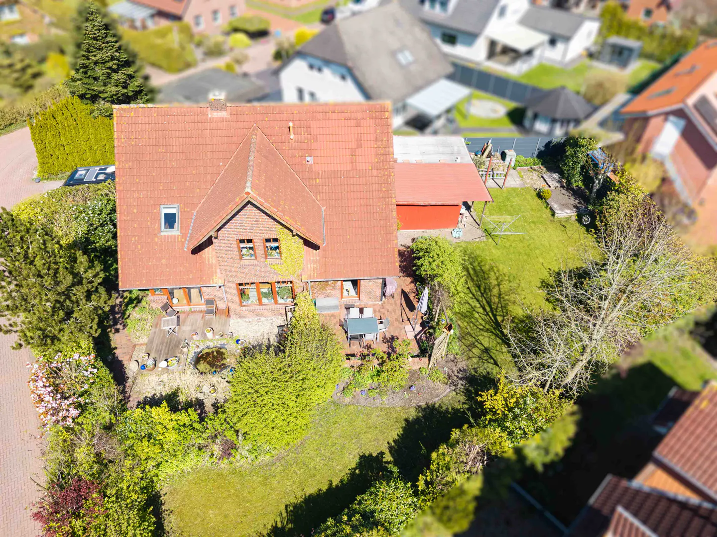 Aerial view of a brick house with a red tile roof, green lawn, and surrounding trees in a residential neighborhood.