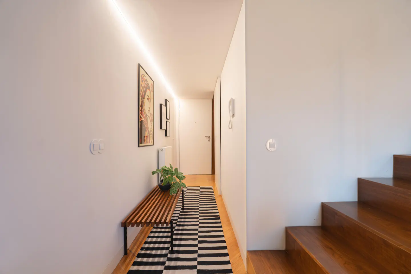 Hallway with white walls, wood floors, and stairs. A striped rug leads to a white door. A bench with a plant sits under framed art.