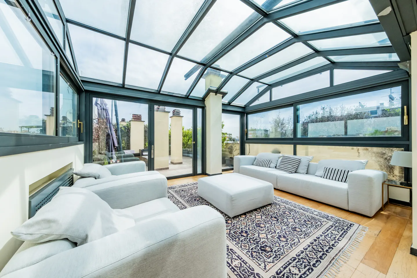 Bright sunroom with glass ceiling, gray frames, and white furniture. A patterned rug sits on the wood floor. Outside, a patio with pillars is visible.
