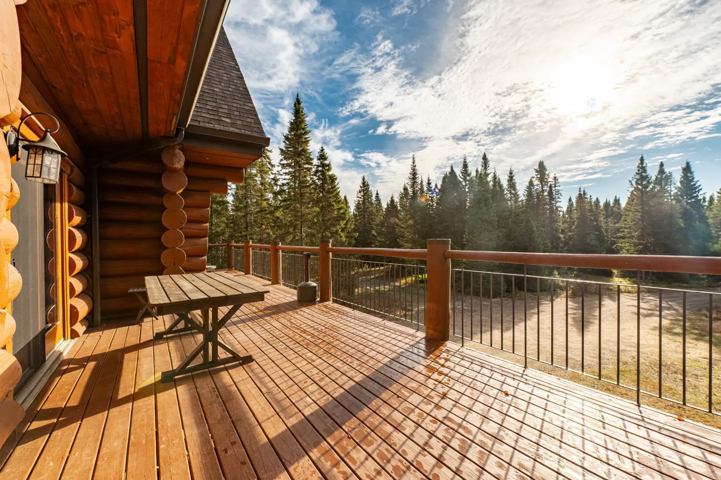 Wooden deck with table and railing overlooking a forest. Log cabin visible on the left. Blue sky with clouds.