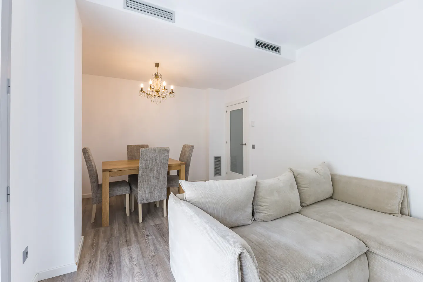 Bright, modern living room with a beige sectional sofa, wood dining table with gray chairs, and a crystal chandelier.