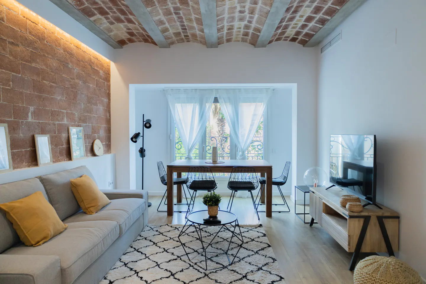 Modern living room with a brick wall, vaulted ceiling, grey sofa with yellow pillows, and a dining area by the window.