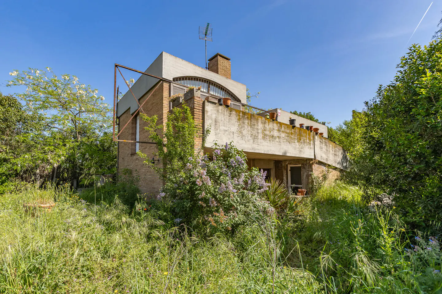 Two-story brick house with a concrete balcony and overgrown yard on a sunny day.