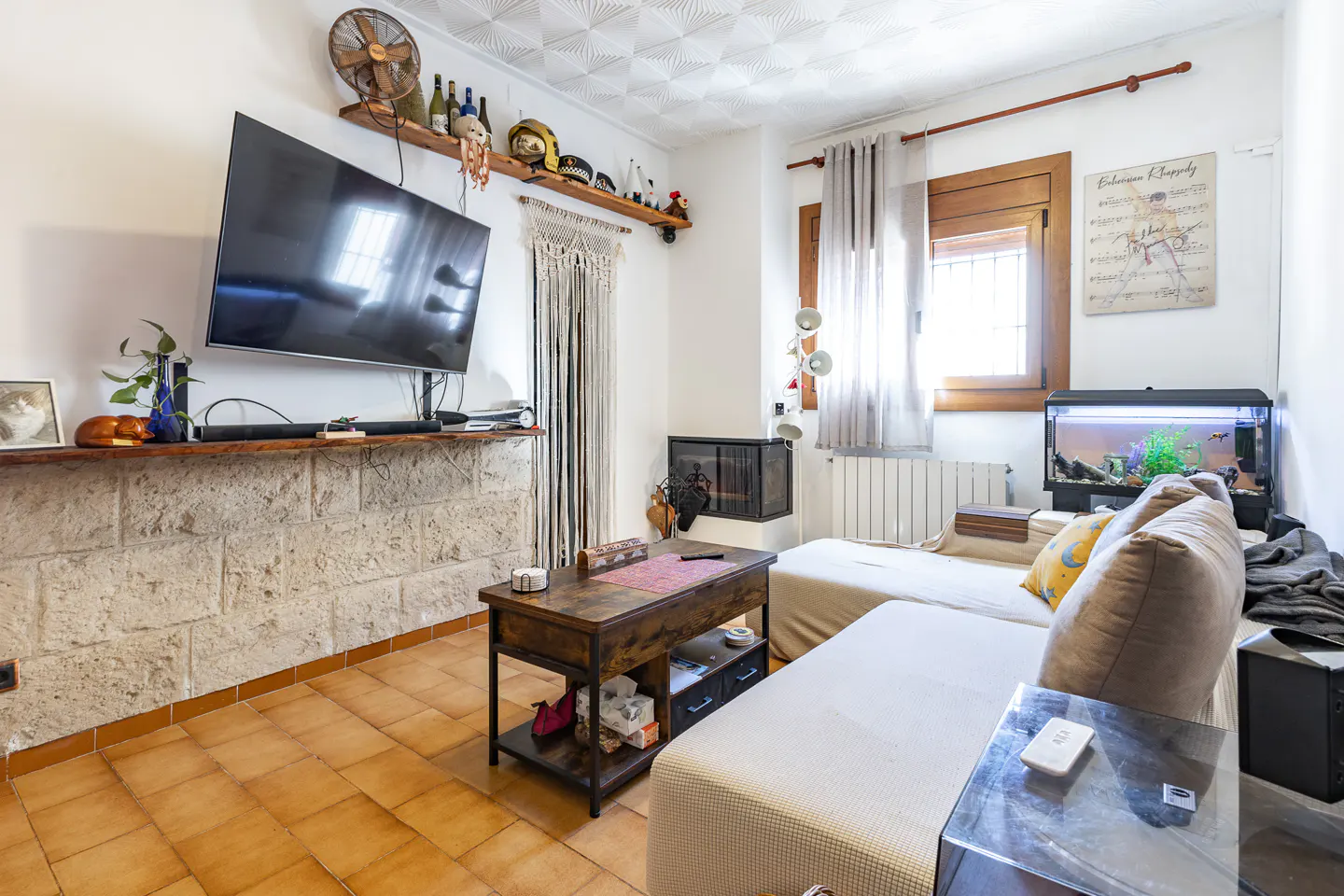 Living room with a TV above a stone shelf, a dark wood coffee table, and a beige sofa. A fish tank sits near a window with sheer curtains.
