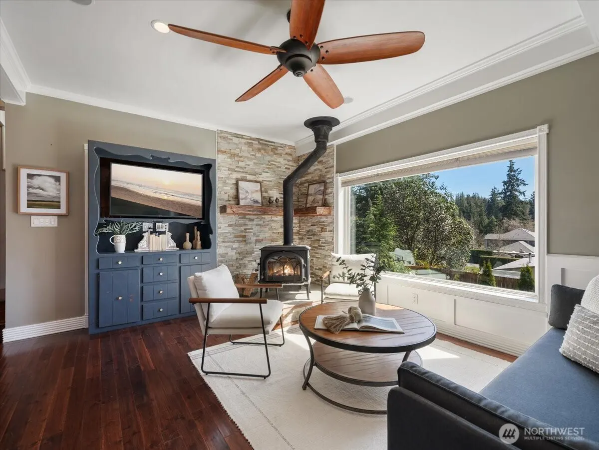 Living room with wood floors, a stone fireplace, and a large window with a view of trees. A blue cabinet holds a TV. A ceiling fan hangs above.