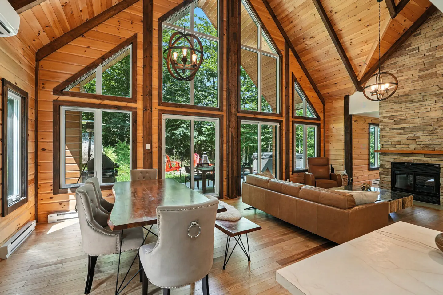 A bright, open-concept living space with wood walls, a stone fireplace, and large windows overlooking a green forest. A dining table and leather sofa are visible.