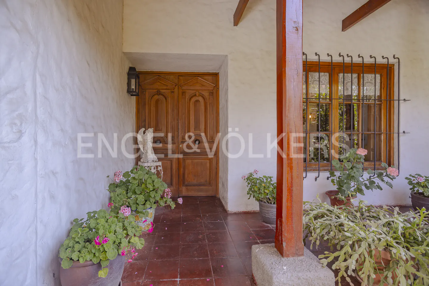 Exterior view of a home with a wooden door, window with bars, and potted plants. The Engel & Völkers logo is visible.