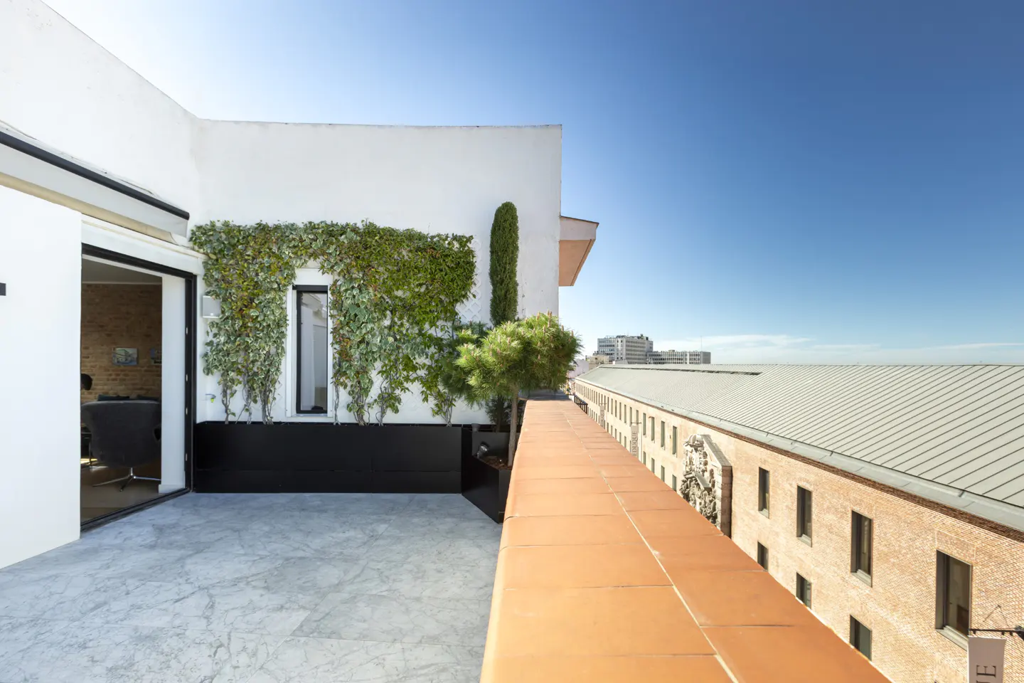 Modern rooftop terrace with marble flooring, a green ivy wall, and a terracotta railing overlooking a brick building.