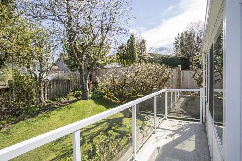 View from a balcony with white railings overlooking a green lawn, trees, and a wooden fence on a sunny day.