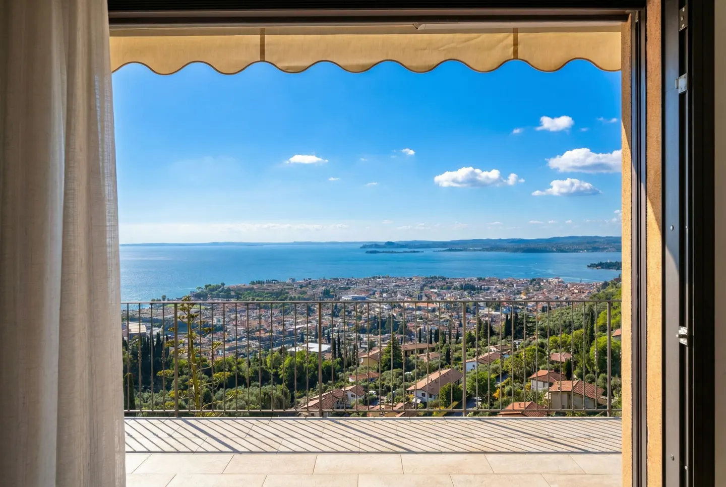 View from a balcony overlooking a city, lake, and blue sky. A tan awning and white curtain frame the scene.