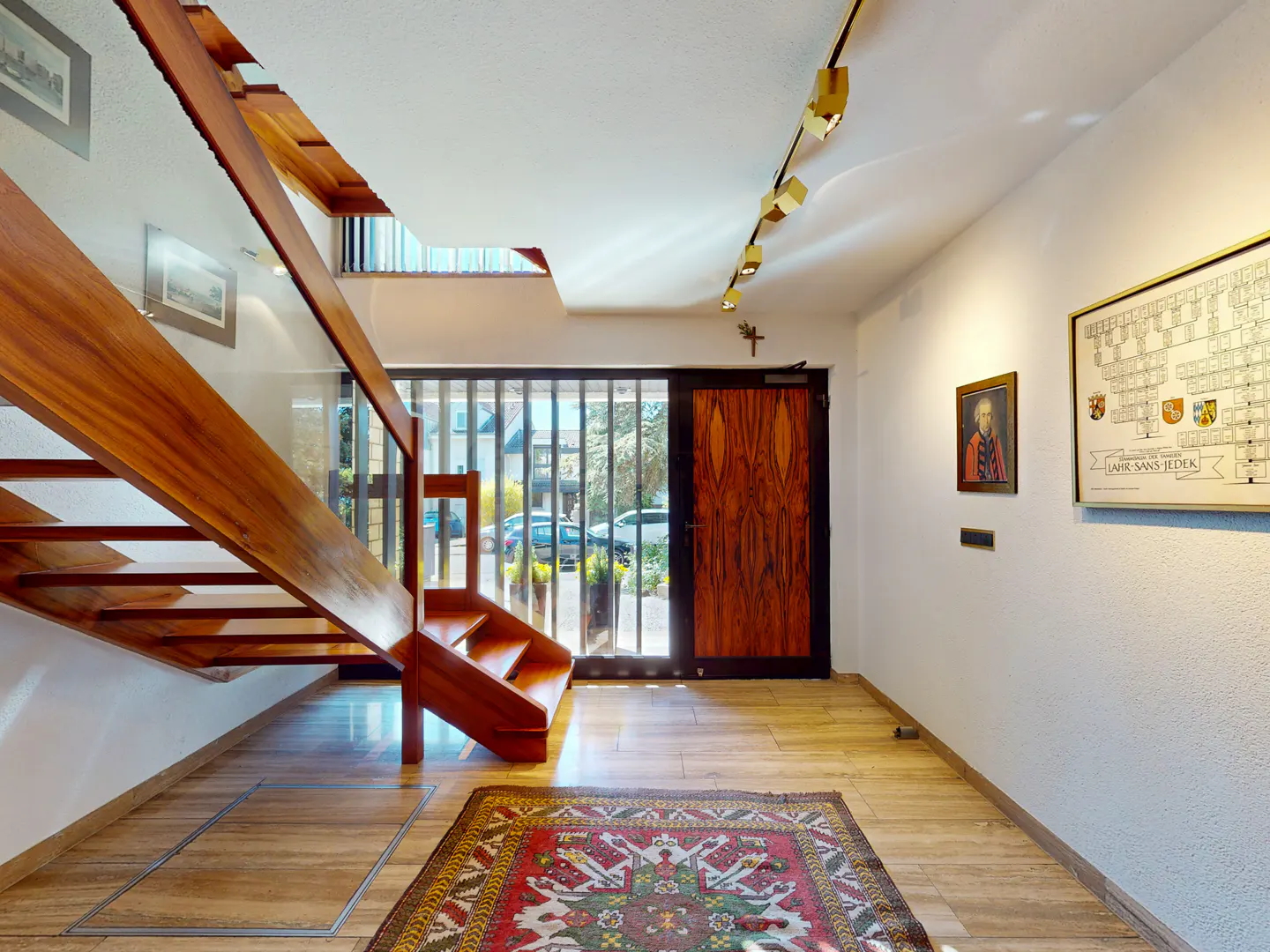 A foyer with a wooden staircase, a patterned rug, and a wooden door. Artwork hangs on the white walls.