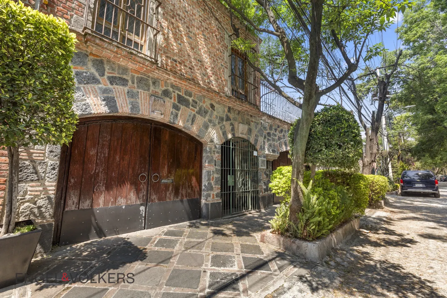 Exterior of a two-story brick and stone house with a large wooden arched door and a wrought iron gate.